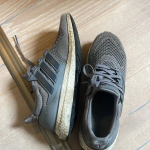 Grå Adidas Ultraboost sneakers - Säljer ett par grå Adidas Ultraboost sneakers med stickad ovandel och klassiska tre ränder på sidan. Skorna har snörning och en vit, bekväm Boost-sula. Perfekta för både träning och vardag.