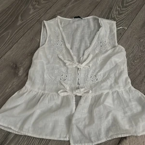 Vit broderad väst med knyt - Söt vit väst med broderade blommor och knyt framtill. Västen har v-ringning och volang nedtill, vilket ger en romantisk känsla. Perfekt att styla över en topp eller klänning för en extra detalj.