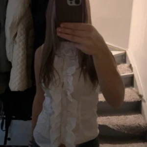 Vit blus med volang från Vero Moda - Säljer en söt vit blus från Vero Moda med volangdetaljer längs framsidan. Blusen är ärmlös och har en feminin och elegant look, perfekt för att lyfta vilken outfit som helst.