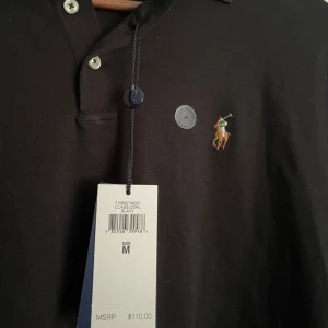 Svart pikétröja från Polo Ralph Lauren - Klassisk svart pikétröja från Polo Ralph Lauren i modellen Classic Fit. Tröjan har korta ärmar, krage med knappar och den ikoniska broderade loggan på bröstet. Perfekt för en stilren och tidlös look. Helt oanvänd med lappar kvar.