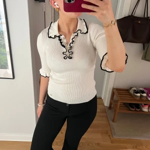 Vit ribbad pikétröja från Holly & Whyte, Lindex - Säljer en vit ribbad pikétröja från Holly & Whyte, Lindex med svarta vågiga detaljer längs krage och ärmslut. Tröjan har lång ärm och figurnära passform, perfekt för en stilren look.