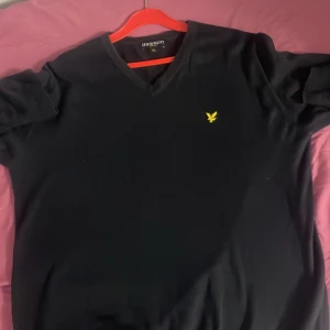Svart v-ringad tröja från Lyle & Scott - Klassisk svart v-ringad tröja från Lyle & Scott med gul broderad logga på bröstet. Tröjan har långa ärmar och är tillverkad i ett mjukt material som passar perfekt till många olika outfits.
