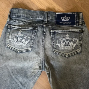 Grå-blå Victoria Beckham x Rock & Republic jeans 25 - Säljer ett par äkta vintage Y2K grå-blå bootcut jeans med snygga broderade kronor på bakfickorna från Victoria Beckham x Rock & Republic. Jeansen har låg midja och klassisk femficksdesign. Korta i längd. Innerbenslängd 70 cm. Ytterbenslängd 90 cm. Midja 25 tum. Designmässiga slitningar. 