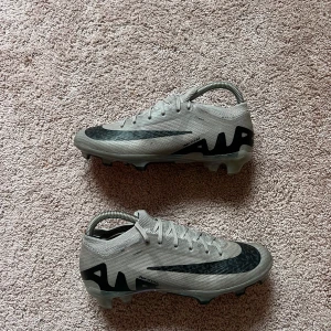 Gråa Nike mercurial 15 Elite - Säljer ett par gråa Nike fotbollsskor med svart swoosh och AIR-tryck på sidan. Skorna har snörning och en mönstrad sula med rosa detaljer undertill. Perfekta för dig som vill sticka ut på planen!