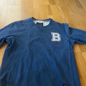 Blå sweatshirt från Bondelid med broderad B - Mörkblå sweatshirt från Bondelid med broderad vit och blå bokstav 'B' på bröstet. Tröjan har rund halsringning och långa ärmar. Perfekt för en avslappnad stil.