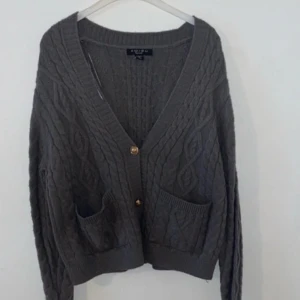 Grå stickad kofta med fickor - Mysig grå stickad kofta från Acne Studios med kabelstickat mönster, två framfickor och knappar framtill. Perfekt för lager-på-lager och har långa ärmar samt djup v-ringning.