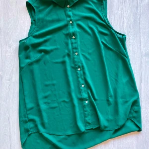 Grön ärmlös skjorta från H&M - Säljer en snygg grön ärmlös skjorta från H&M. Skjortan har klassisk krage och knappar framtill. Tillverkad i mjuk och lätt polyester, perfekt för varmare dagar eller lager på lager.