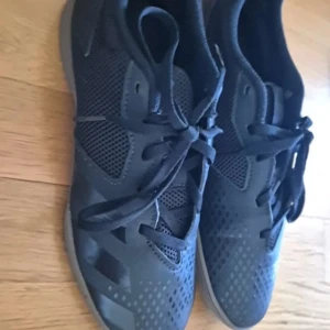 Svarta Adidas sneakers med meshdetaljer - Säljer ett par svarta Adidas sneakers med klassiska ränder på sidan och meshpaneler för extra ventilation. Skorna har snörning och platt sula, perfekt för träning eller vardagsbruk. Snygg och sportig design med diskreta detaljer.