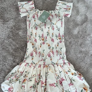 Blommig kortklänning med volang från hm - Supersöt kortklänning med blommigt mönster i rosa, grönt och gult. Klänningen har korta ärmar, smockad överdel och volang nertill. Perfekt för sommarens alla tillfällen! (Inte min bild) ❤️