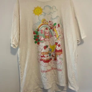 Vit Strawberry Shortcake t-shirt med tryck - Söt vit t-shirt med färgglatt Strawberry Shortcake-tryck på ryggen. Motivet har jordgubbar, regnbåge och figurer i pastellfärger. Tillverkad i 100% bomull och har en avslappnad passform med korta ärmar. Köpt på pacsun i LA! 