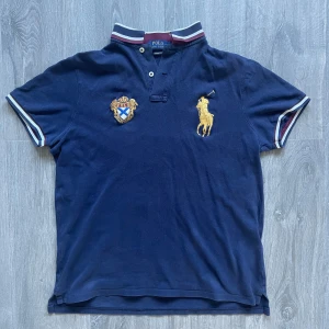 Marinblå piké från Polo Ralph Lauren - Säljer en marinblå pikétröja från Polo Ralph Lauren med broderad gul polospelare och ett snyggt vapensköldsbroderi på bröstet. Tröjan har korta ärmar och vit-röd randig krage. Custom slim fit-modell.