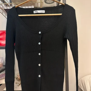 Svart ribbad kofta från ZARA - Säljer en svart ribbad kofta från ZARA med lång ärm och knappar framtill. Koftan har en rundad ringning och är tillverkad i ett stretchigt material som sitter snyggt på kroppen. Passar även mig som är en storlek S! Buren en gång.