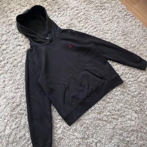 Svart hoodie från Polo Ralph Lauren - Säljer en svart hoodie från Polo Ralph Lauren med klassisk röd broderad logga på bröstet. Tröjan har lång ärm, huva och en stor magficka framtill. Perfekt för en avslappnad och stilren look.