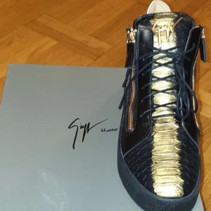 Svarta och guldiga sneakers från Giuseppe Zanotti - Unika sneakers i svart och guld med reptilmönstrad textur, snörning och dragkedjor på sidorna. Skorna har rund tå och platt sula, samt exklusiva gulddetaljer på plösen och sidan.