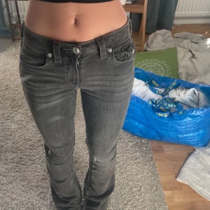 Grå jeansbyxor från True Religion - Säljer ett par grå jeansbyxor från True Religion med klassiska detaljer och synliga sömmar. Byxorna har låg midja, raka ben och silverfärgade knappar. Snygg passform och ikonisk bakficka med lock och knapp.