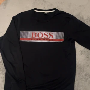 Svart långärmad tröja från Hugo Boss - Säljer en svart långärmad tröja från Hugo Boss med stort BOSS-tryck i rött och vitt framtill. Tröjan har rund halsringning och en stilren design som passar perfekt till vardags. Klassisk och enkel modell.