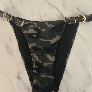 Camo bikiniunderdel med spännen - Säljer en bikiniunderdel i camomönster med justerbara spännen i sidorna. Färgerna är svart, grå och grön. Modellen har en normal passform och är tillverkad i syntetmaterial. Perfekt för sommaren!