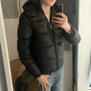 Svart dunjacka från Polo Ralph Lauren - Säljer en svart dunjacka från Polo Ralph Lauren med huva och dragkedja. Jackan har en röd broderad logga på bröstet och flera praktiska fickor med dragkedja. Perfekt för kyliga dagar och riktigt snygg passform. Den är i extremt fint skick, finns inga skavanker eller dyligt på jackan. Finns orderbekräftelse men inget kvitto tyvärr, den är äkta (kolla 5:e bilden) hör av er vid frågor!🤩