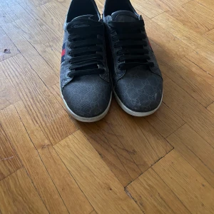Gucci sneakers med monogram och röd/blå detalj - Säljer ett par svarta Gucci sneakers med klassiskt GG-monogram, svarta skosnören och röd/blå rand på sidan. Baksidan har metallicröda och lila detaljer med Gucci-logga. Perfekta för dig som vill sticka ut med stil.