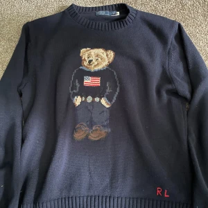 Mörkblå stickad tröja med björn från Polo Ralph Lauren - Mörkblå stickad tröja med rund halsringning och ikonisk broderad björn på bröstet. Björnen har en tröja med amerikansk flagga och jeans. RL-broderi i rött nedtill. 
