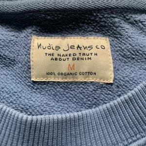 Blå sweatshirt från Nudie Jeans - Nypris: ~180€.                                            Säljer en blå sweatshirt från Nudie Jeans i 100% ekologisk bomull. Tröjan har rund halsringning, långa ärmar och en broderad detalj med ett glas på bröstet. Perfekt för en avslappnad stil.