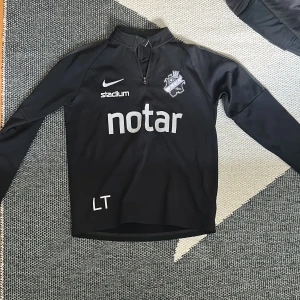 Svart Nike Dri-Fit AIK träningströja - Svart långärmad träningströja från Nike med AIK-logga på bröstet och sponsorer tryckta fram och bak. Tröjan har halv dragkedja framtill och är tillverkad i Dri-Fit material som andas. Perfekt för fotbollsträning.