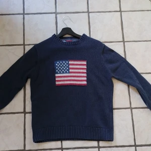 Mörkblå stickad tröja från GANT med amerikansk flagga - Mörkblå stickad tröja från GANT i 100% bomull. Tröjan har en stor amerikansk flagga broderad på bröstet och ribbade muddar vid ärmslut och nederkant. Klassisk och stilren design med GANT-logga på axeln. 