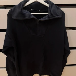 Svart stickad tröja med dragkedja - Mysig marinblå stickad tröja med hög krage och dragkedja framtill. Tröjan har långa ärmar och ribbade detaljer vid krage, ärmslut och nederkant. Perfekt för kyligare dagar och enkel att styla till olika outfits.