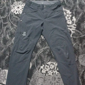 Svarta Haglöfs byxor - Svarta friluftsbyxor från Haglöfs med diskret logga på låret. Byxorna har dragkedja och knapp i midjan samt förstärkta partier vid knäna. Perfekta för utomhusaktiviteter och äventyr.