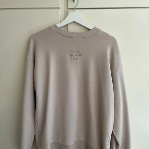 Beige stickad tröja från New Garments - Säljer en stilren beige stickad tröja från New Garments. Tröjan har rund halsringning, diskret broderad text framtill och långa ärmar. Perfekt för lager-på-lager och enkel att matcha med olika outfits.