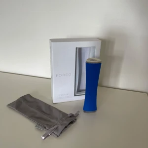Blå Foreo Espada ansiktsbehandling - En blå Foreo Espada, ett elektriskt verktyg för ansiktsbehandling med LED-ljus. Kommer med originalförpackning och en grå förvaringspåse i tyg. Perfekt för hudvård och punktbehandling av orenheter.