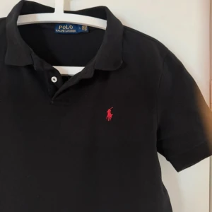 Svart pikétröja från Polo Ralph Lauren - Klassisk svart pikétröja från Polo Ralph Lauren med röd broderad logga på bröstet. Tröjan har korta ärmar, krage och knäppning med två knappar. Perfekt för en stilren och avslappnad look.