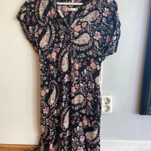 Blommig svart klänning från Bronco Jeans - Svart klänning med blommigt mönster i rosa, grönt och vitt från Bronco Jeans. Klänningen har korta ärmar, knappar framtill och ett knytband i midjan för en fin passform. Perfekt för vår och sommar! Storlek M men passar även S