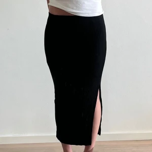 Svart ribbad långkjol från NA-KD - Säljer en svart ribbad långkjol från NA-KD i storlek XS. Kjolen har en tight passform och en snygg slits på sidan som ger en stilren look. Säljer för den ej är min stil!