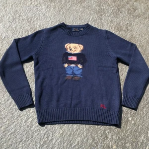 Mörkblå stickad tröja med björn från Polo Ralph Lauren - Mörkblå stickad tröja från Polo Ralph Lauren med ikonisk björn på bröstet. Björnen har en tröja med amerikansk flagga. Rund halsringning och ribbade muddar vid ärmslut och nederkant. Klassisk och tidlös design. Perfekt för dig som letar något fräscht till sommaren!☀️ Storlek herr S