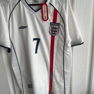 England fotbollströja Beckham 7 - Säljer en vit England fotbollströja från Umbro med nummer 7 och Beckham på ryggen. Tröjan har korta ärmar, röd vertikal rand och Englands landslagsmärke på bröstet. Perfekt för fans av landslaget och klassiska spelare.