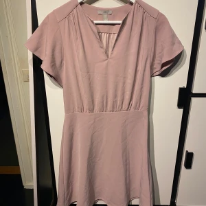 Ljusrosa kortklänning från H&M - Säljer en söt ljusrosa kortklänning från H&M i storlek 36. Klänningen har v-ringning, korta ärmar och en lätt utställd kjol. Perfekt för vår och sommar! Materialet känns mjukt och lätt mot huden.