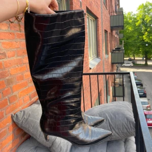 Svarta höga stövlar med krokodilmönster NAKD - Säljer ett par svarta höga stövlar med krokodilmönster och spetsig tå. Köpt på NAKD för 900kr, sparsamt använda. Skorna har klack och är tillverkade i glansigt material. Perfekta för dig som vill sticka ut med en edgy look! Obs hälen är lite nedgången.