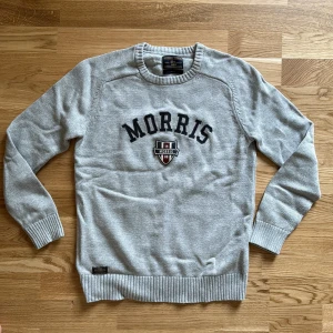 Grå stickad tröja från Morris Gents - Säljer en ljusgrå stickad tröja från Morris Gents med broderad logotyp och text på bröstet. Tröjan har rund halsringning, långa ärmar och ribbade muddar vid ärmslut och nederkant. Perfekt för en stilren och avslappnad look.