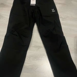 Svarta byxor från Haglöfs Buteo Pant Women - Svarta byxor från Haglöfs, modell Buteo Pant Women. Byxorna har elastisk midja och är tillverkade i ett lätt syntetmaterial, perfekta för utomhusaktiviteter. Diskret Haglöfs-logga på benet och meshfoder på insidan.