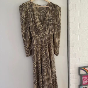 Adoore klänning zebra - Adoore långklänning med zebramönster i beige och mörkbrunt. Klänningen har v-ringning, långa ärmar och ett knytband i midjan. Stl 40