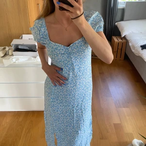 Blå blommig klänning från Chiquelle - Blå klänning med vitt blommönster från Chiquelle som tyvärr inte finns längre. Klänningen har kort puffärm, går till mitten på vaderna samt med en slits på ena benet. Storlek 38, aldrig använd pga lite för stor för mig. Nypris 899kr