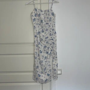Vit blommig sommarklänning  - Söt vit klänning med blått blommönster och smala axelband. Klänningen har rynkdetalj och knyt framtill samt en figurnära passform. Perfekt för varma dagar och midsommarfirande.
