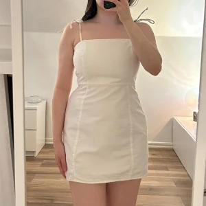 Vit kort klänning med smala axelband - Säljer en stilren vit kortklänning med smala axelband som knyts på axlarna. Klänningen har rak halsringning och dragkedja på ena sidan. Materialet påminner om jeans. Storlek saknas, men skulle uppskatta att det är en XS.