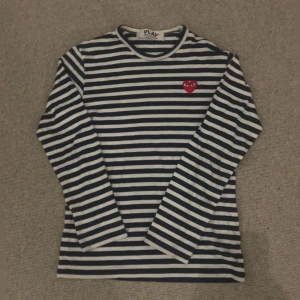 Randig långärmad tröja från Comme des Garçons Play - Säljer en klassisk långärmad tröja från Comme des Garçons Play med marinblå och vita ränder. Tröjan har ett rött hjärta med ögon broderat på bröstet. Perfekt för en stilren och trendig look.