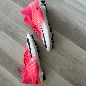 Nike Mercurial rosa fotbollsskor - Snygga Nike Mercurial fotbollsskor i rosa och vitt med svarta detaljer. Skorna har hög strumpliknande krage, snörning och mönstrad ovansida. Perfekta för dig som vill sticka ut på planen och få grym kontroll på bollen.