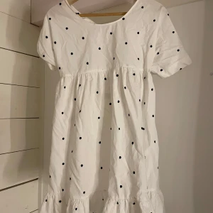 Vit prickig kortklänning med öppen rygg - Supersöt vit kortklänning med svarta prickar, volangkant nertill och korta ärmar. Klänningen har öppen rygg med knytband upptill. Perfekt för en somrig look!! 💗☀️ Endast använd en gång! 