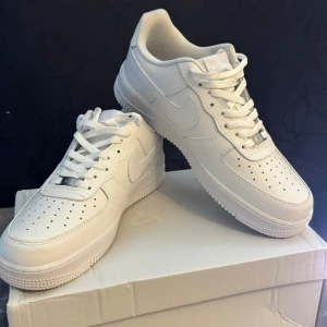 Nike Air Force 1 - Klassiska Nike Air Force 1 i helvitt skinn med perforerad tå, vit sula och ikonisk swoosh på sidorna. Lågt skaft, snörning och stilren look som alltid funkar. Perfekta för dig som vill ha en clean och tidlös sneaker. Storlek 43                                                             Aldrig använda!