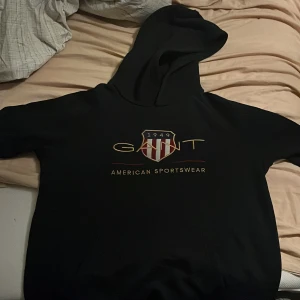Svart hoodie från GANT med tryck hål på luvan dock därför priset - Svart hoodie från GANT med stort tryck på bröstet och klassisk huva. Framsidan har GANT-logga och ett sköldmärke med röda och vita detaljer. Mjuk och skön, perfekt för chill dagar. Snygg och enkel att matcha med jeans eller joggers.