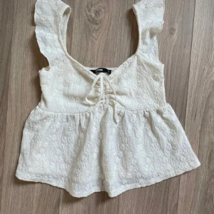 Vit volangtopp från BikBok XS - Supersöt vit topp från BikBok i storlek XS. Modellen har volangärmar, knytning framtill och en lätt strukturerad yta. Perfekt för dig som gillar romantisk och luftig stil. Passar extra bra till sommarens outfits!